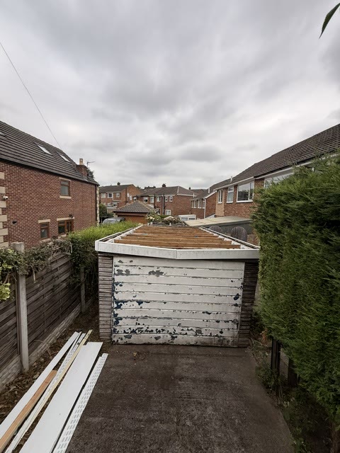 Garage with flaking white up-and-over door and a rotten timber flat roof
